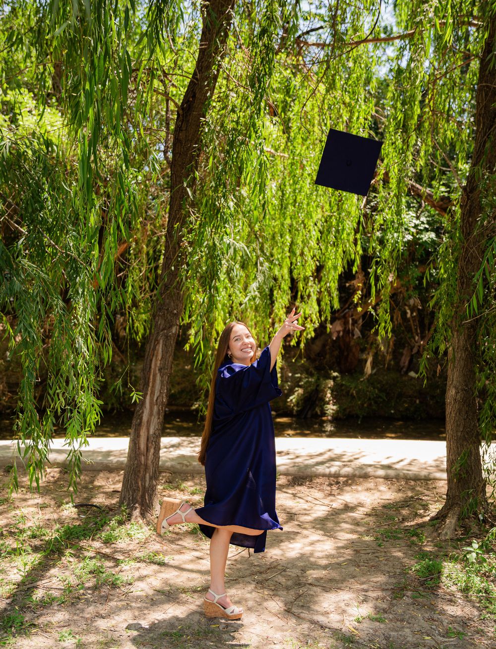 Happy graduate in navy cap and gown tossing mortarboard under willow trees.