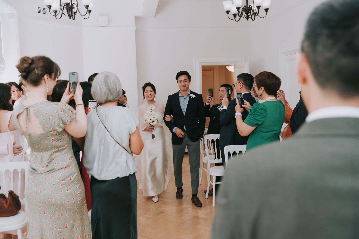 Couple walks down aisle at elegant indoor wedding ceremony while guests photograph and celebrate the special moment.