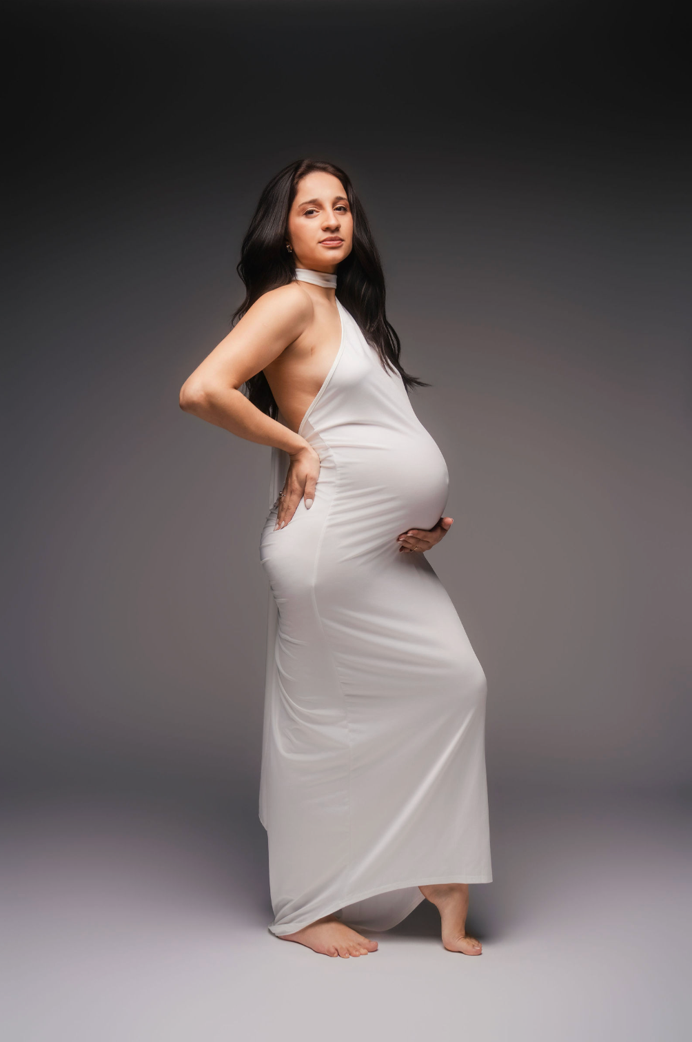 Pregnant woman in white dress holding baby bump, side profile shot on gray background.