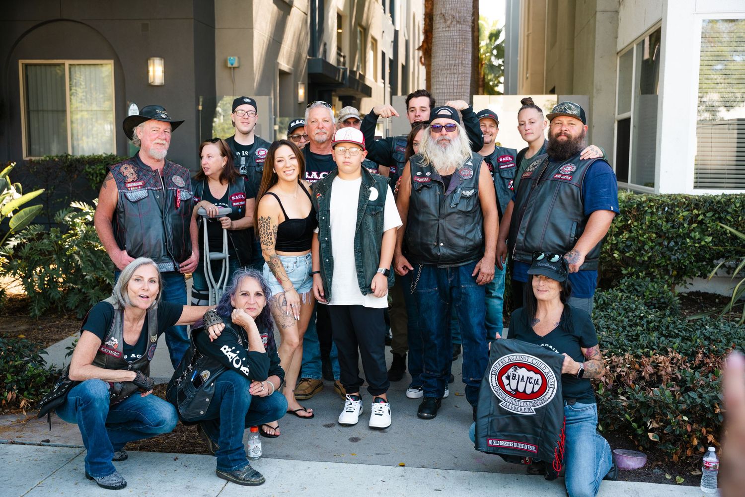Large group of motorcycle club members posing together outdoors near a building for a group photo.