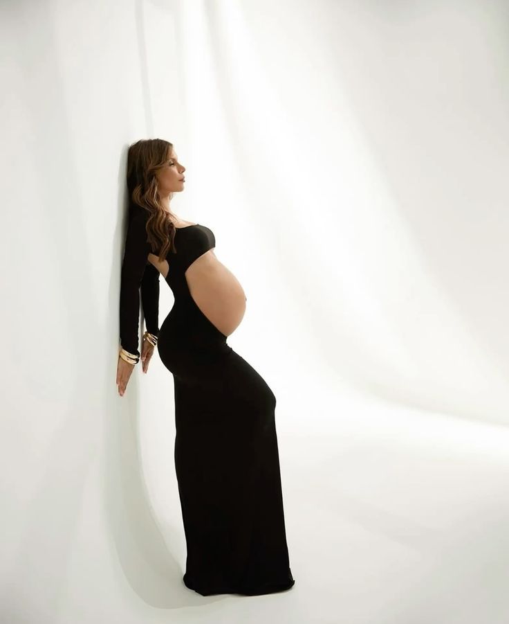 Pregnant woman in black dress posing on a sunny beach with ocean waves, showcasing her baby bump.