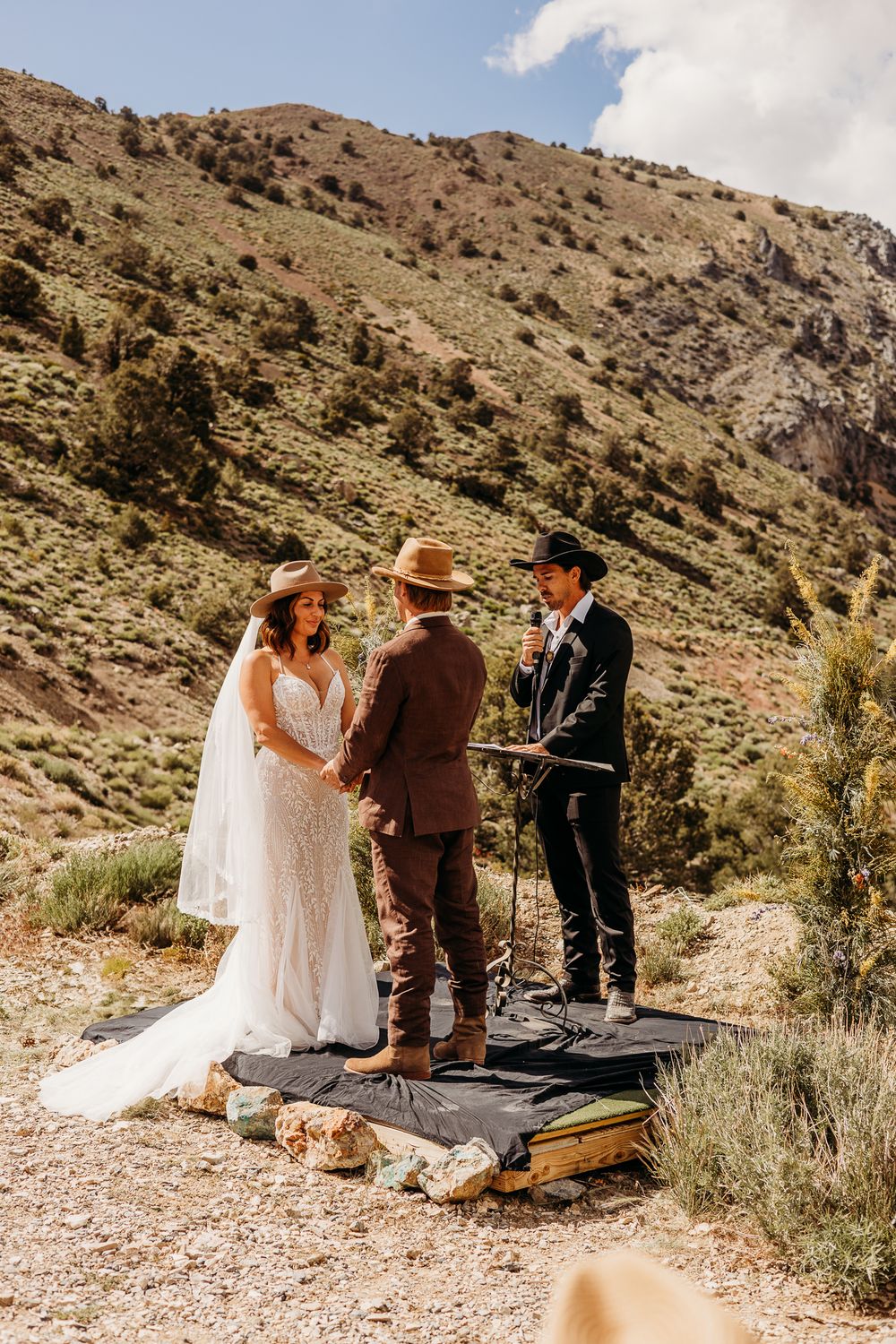 Outdoor wedding ceremony in mountain setting with bride in white gown, groom in brown suit, both wearing cowboy hats.