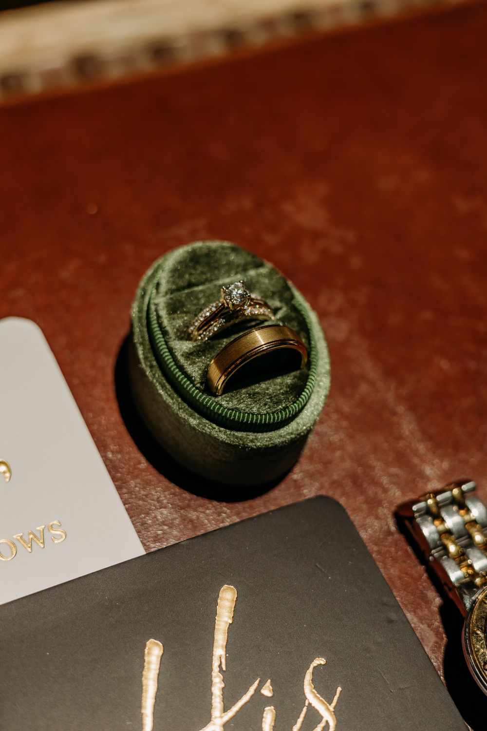 Gold wedding rings displayed in a green velvet oval ring box on a dark surface with elegant cards nearby.