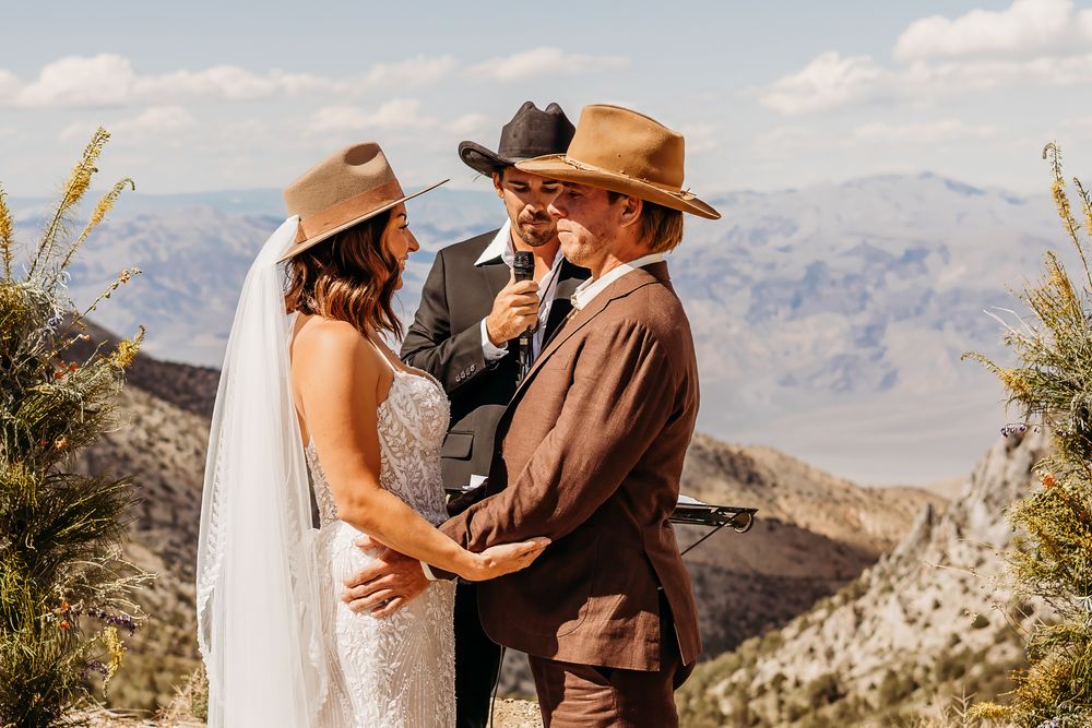 Bride and groom exchange vows outdoors with mountain views, officiant in cowboy hat, rustic western wedding ceremony.
