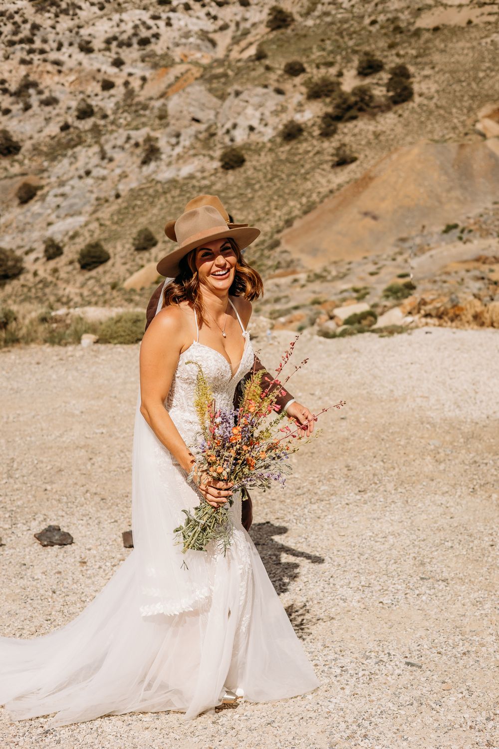 Smiling bride in white mermaid gown, tan cowboy hat, holding wildflower bouquet in desert landscape.