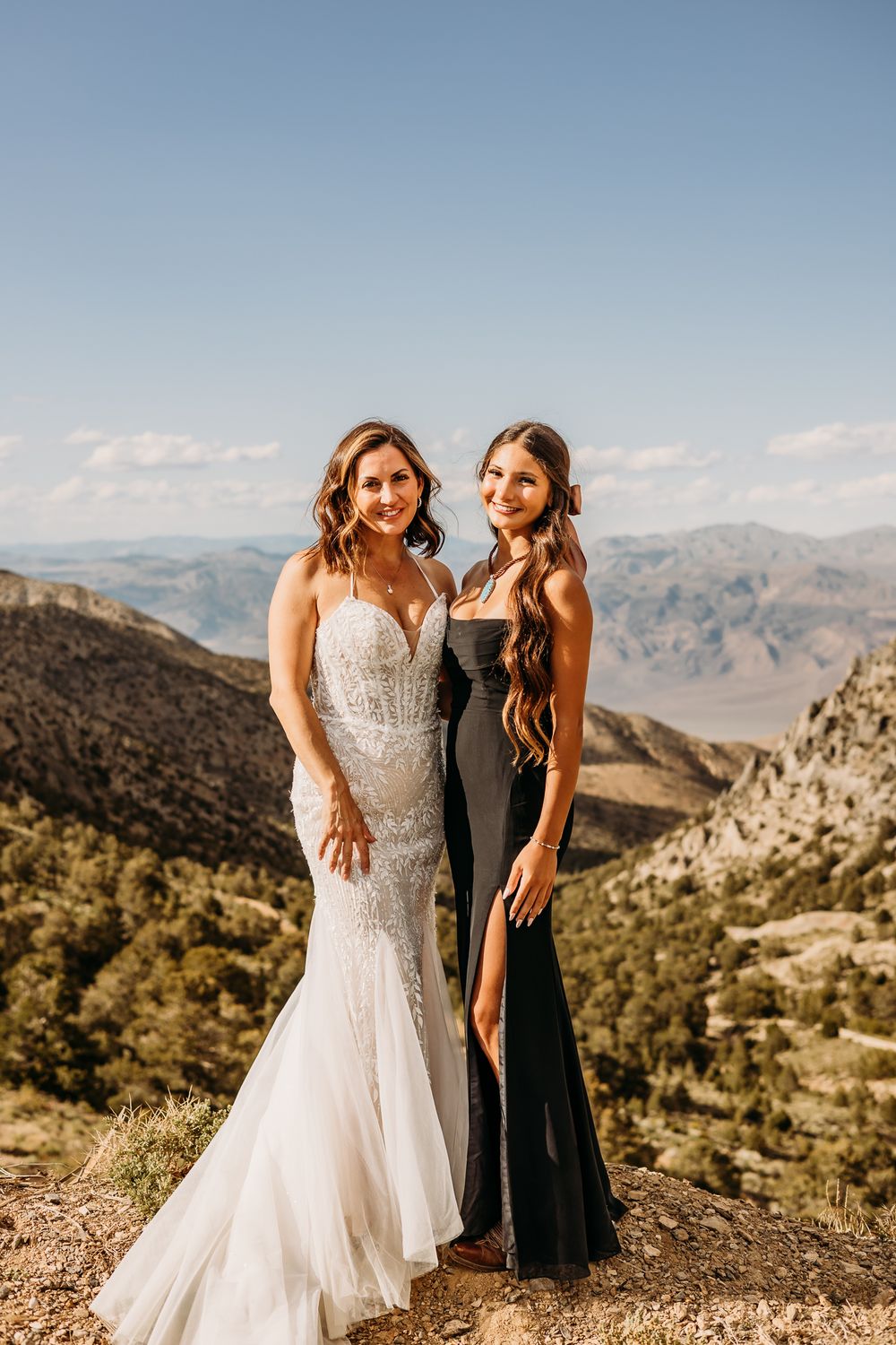 Bride in white gown and bridesmaid in black dress posing together on a mountain overlook under clear blue sky.