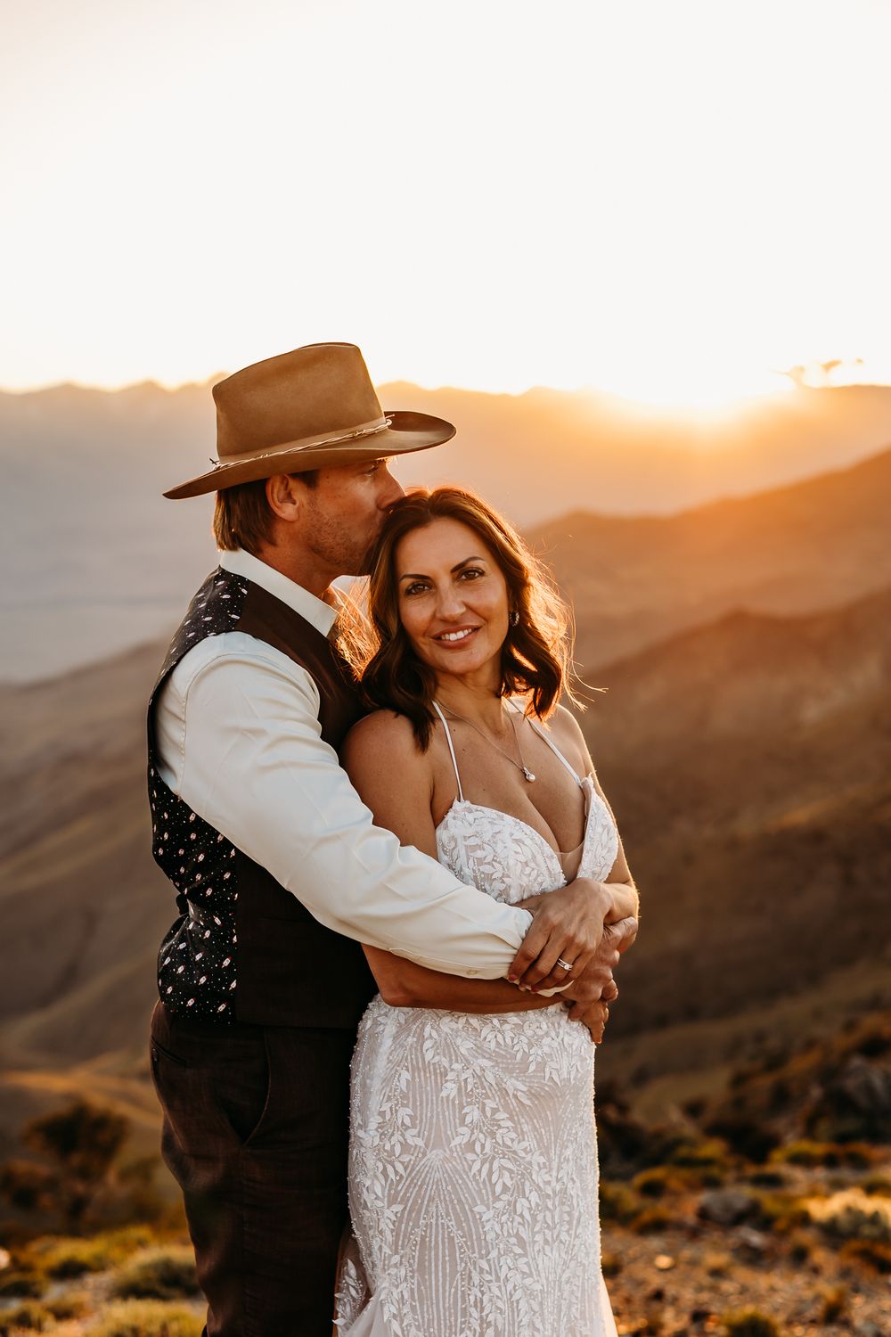 Couple embracing at golden sunset on mountain top, bride in lace gown, groom in vest and wide-brim hat.