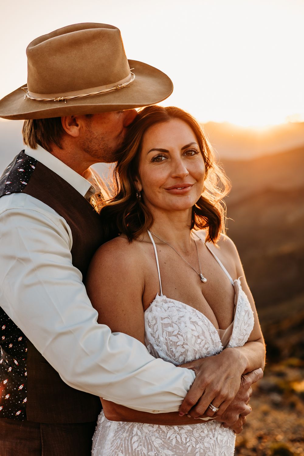 Bride and groom embrace at golden hour sunset, groom in cowboy hat kissing bride's cheek in romantic outdoor wedding portrait.