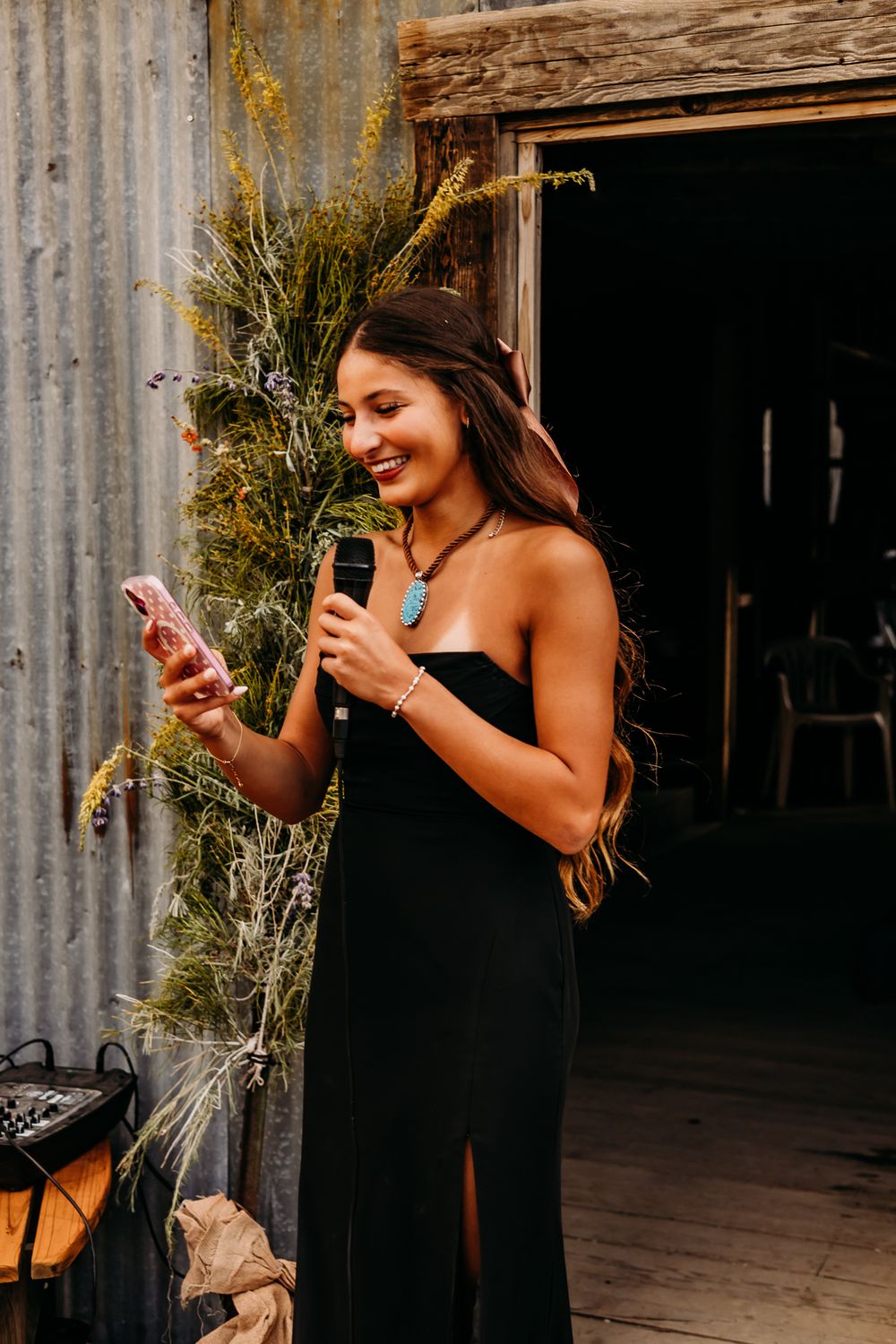 Smiling woman in black strapless dress holding microphone and phone at rustic barn event.