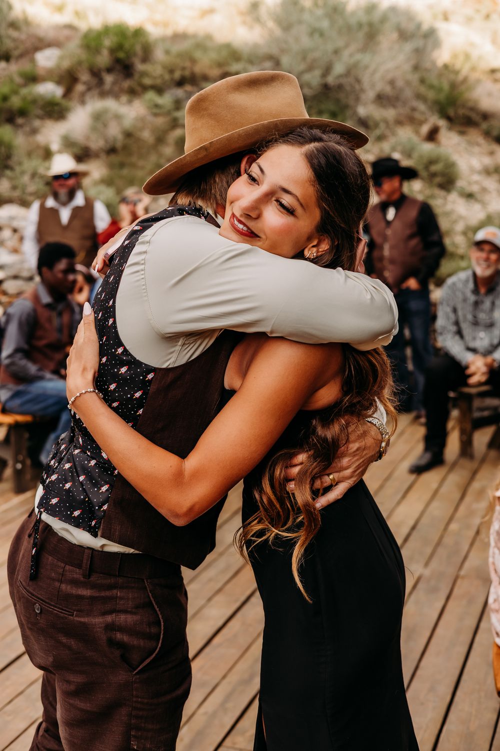Two people share a warm hug at a rustic outdoor western-themed wedding ceremony on a wooden deck.