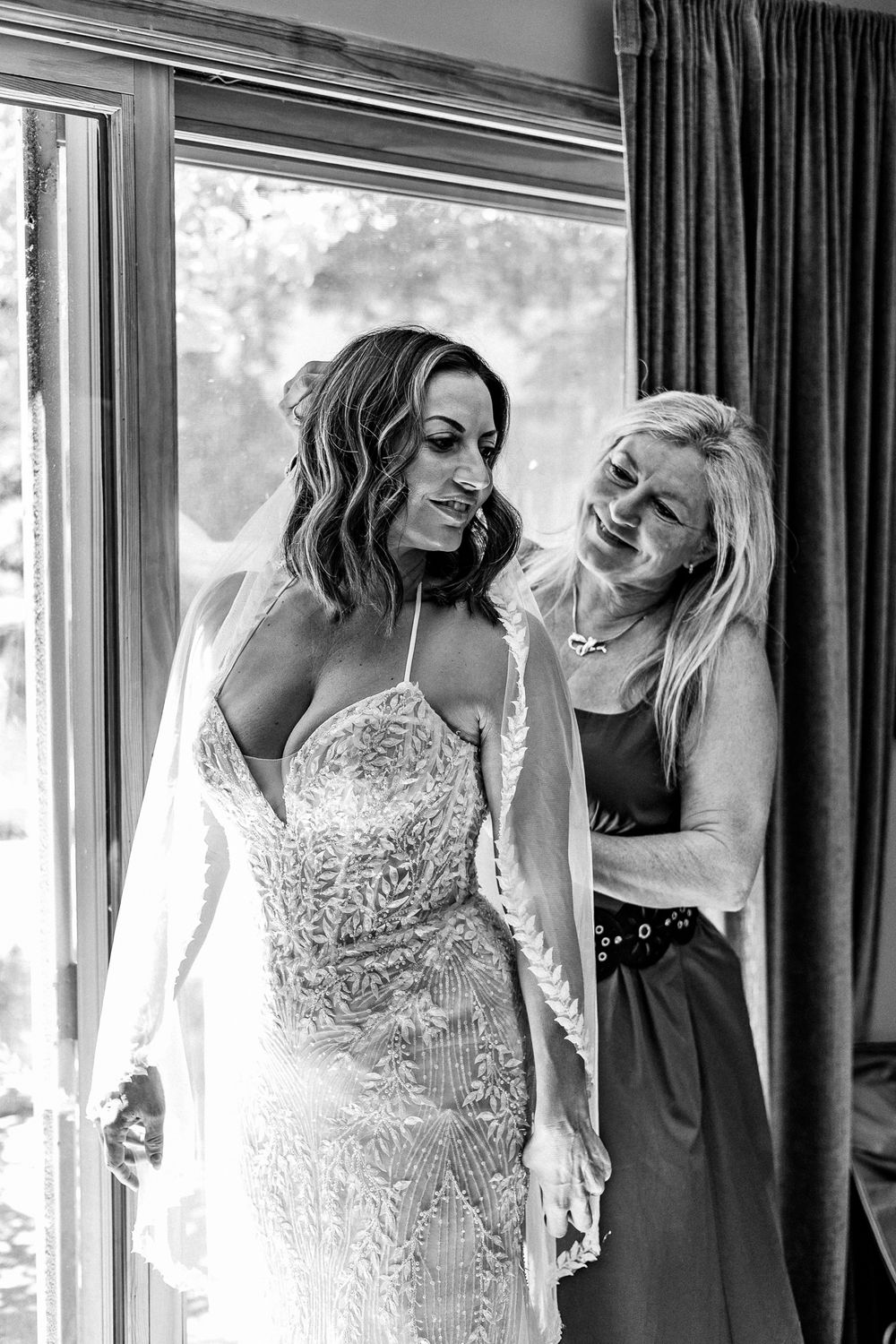 Black and white photo of a bride in a beaded gown smiling at her mom helping her prepare near a window.