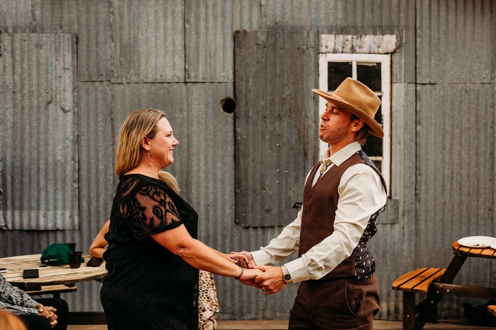Groom in a cowboy hat and vest holds hands with his sister in a black dress outdoors near a rustic metal building.