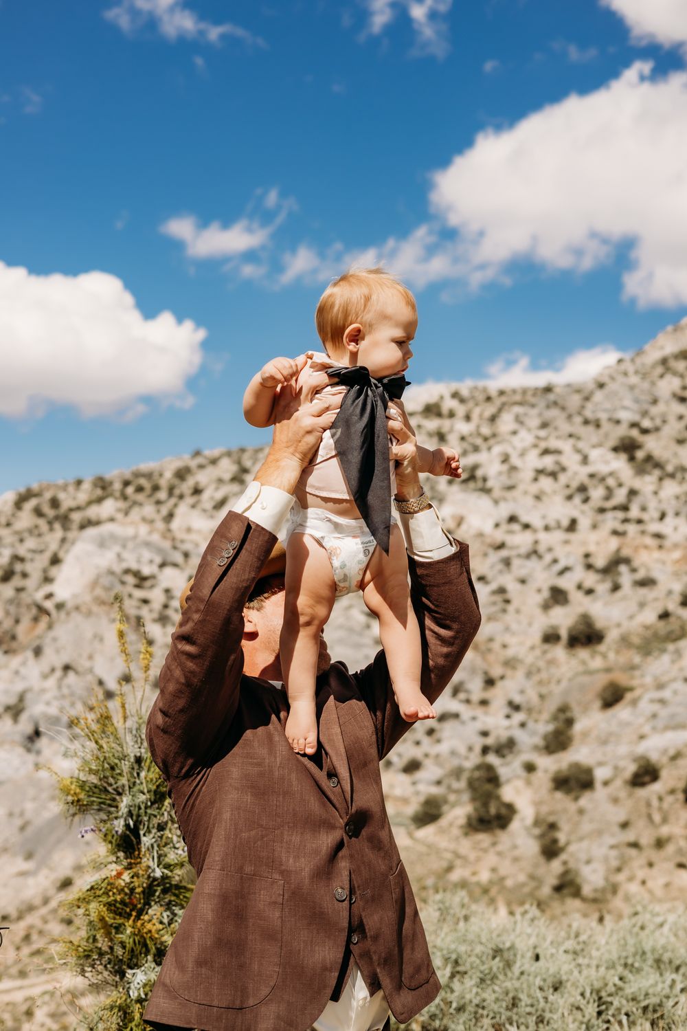 Father in brown suit lifts smiling baby wearing black bow tie against rocky mountain landscape and blue sky.