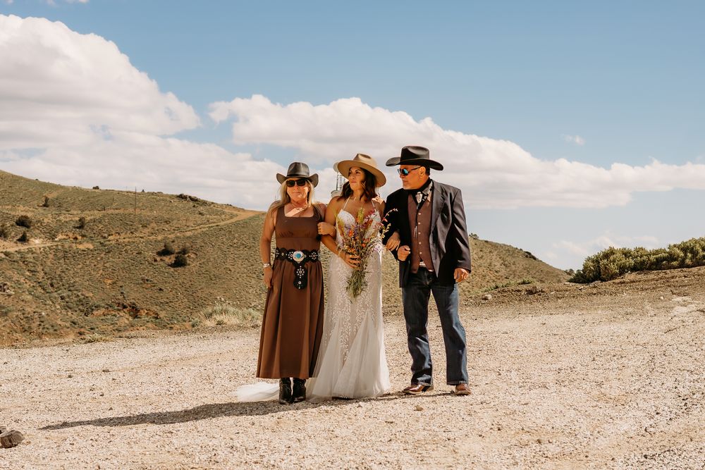 Bride in white gown walks with her parents in cowboy hats on a dusty desert path under blue skies.