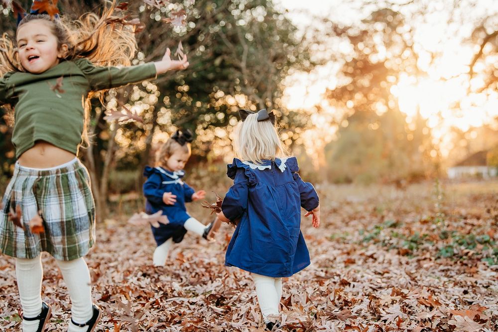 Three young girls playing and running through fallen autumn leaves at golden sunset in park.
