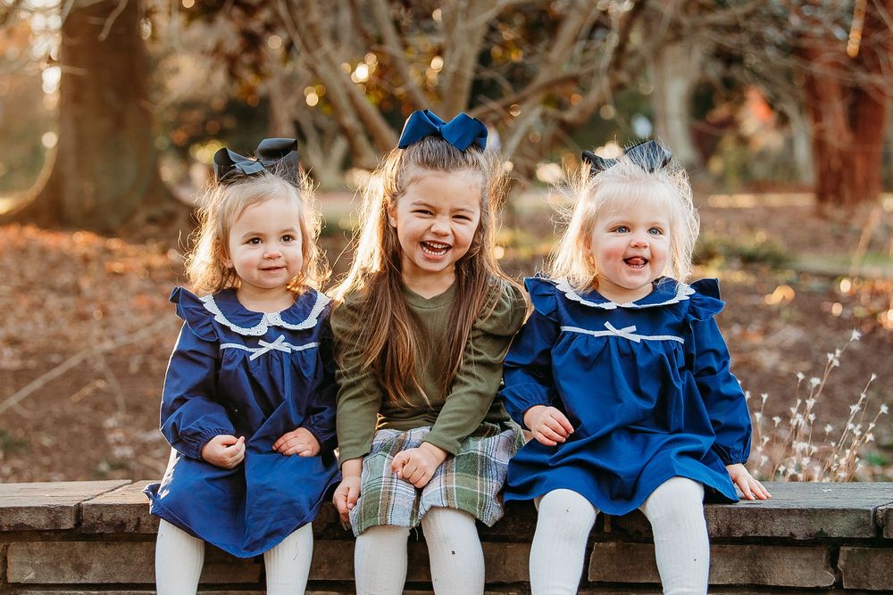 Three young girls in matching navy blue dresses with bows sitting on stone bench outdoors.