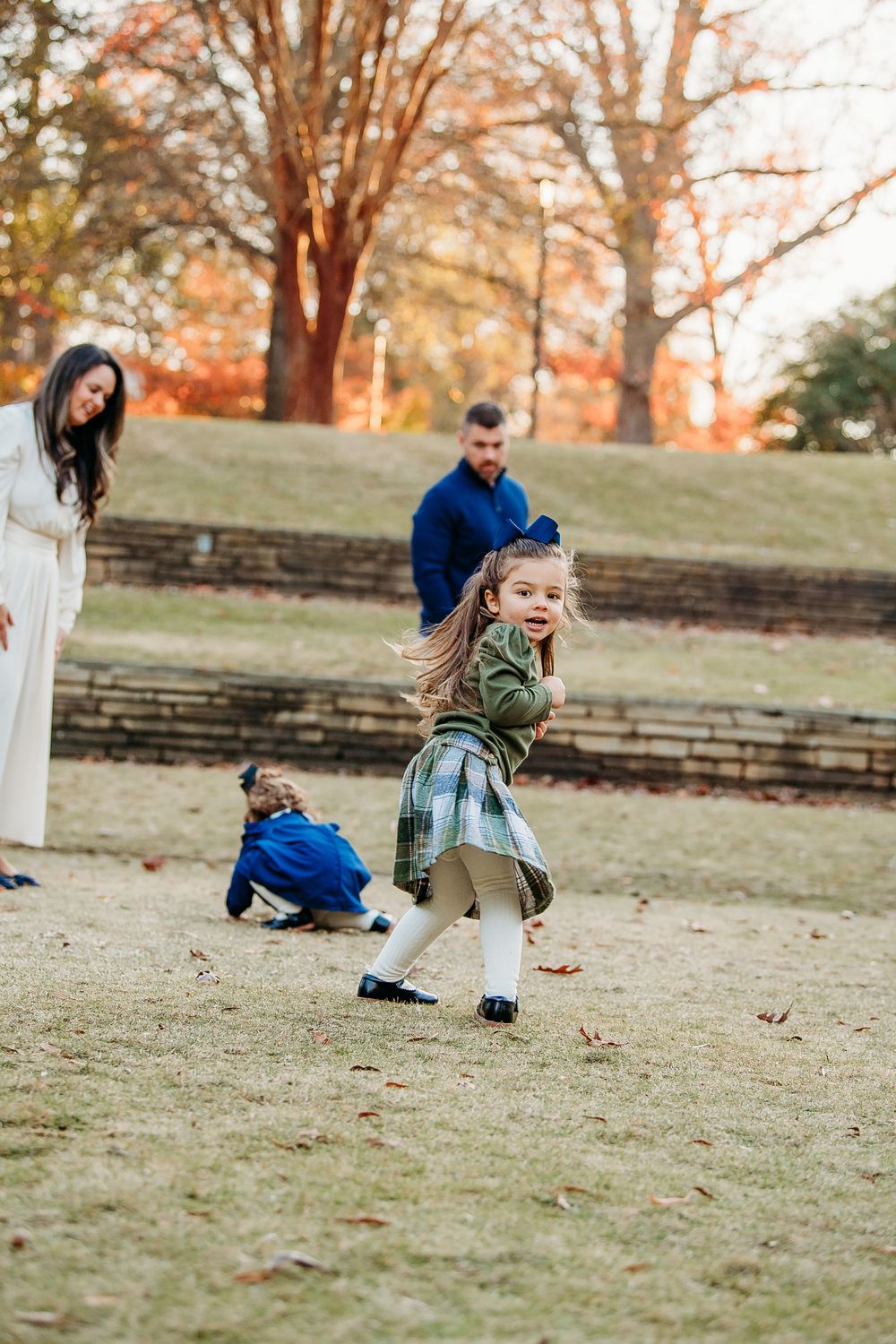 Young girl in plaid skirt running playfully on grass while family watches in autumn park.