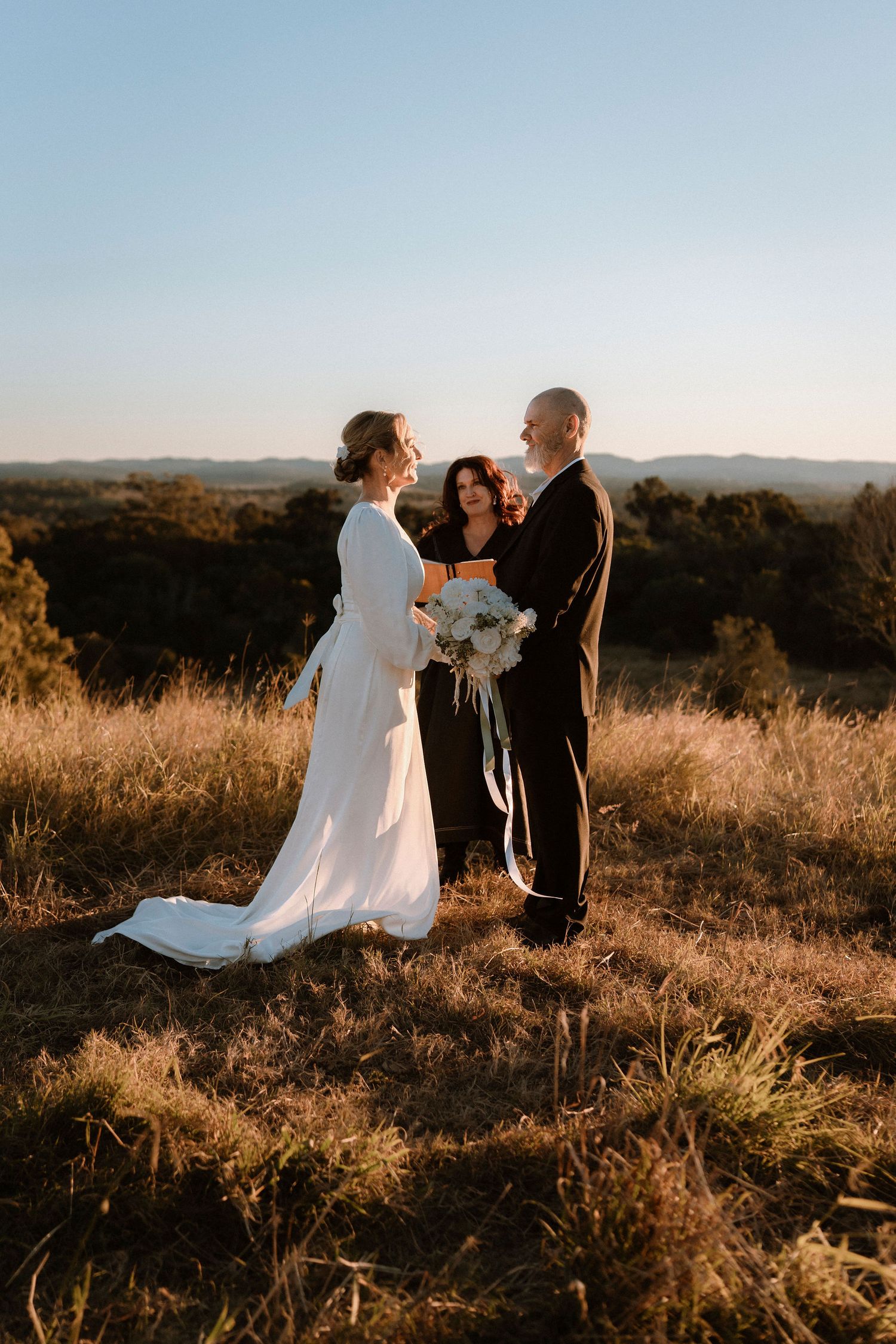 Bride and groom exchange vows with officiant on golden hilltop at sunset, bride holding white bouquet.