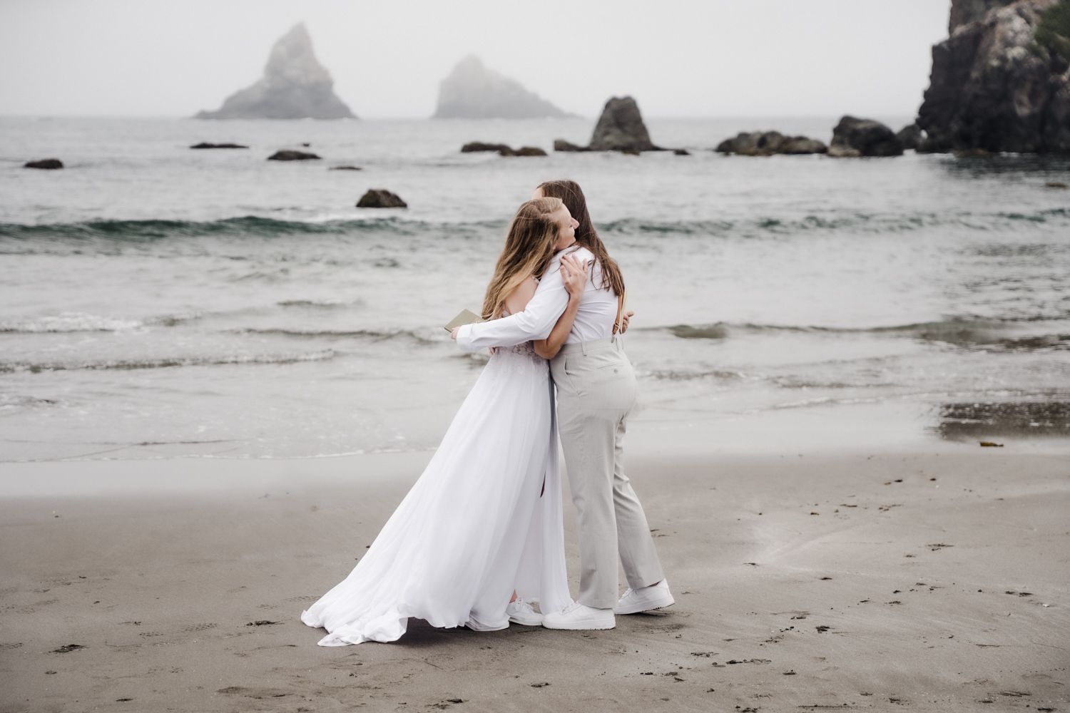 Two brides in white wedding gowns embrace on a misty beach with rocky sea stacks in the background.