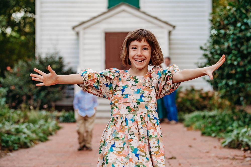 A joyful young girl in a floral dress spreads her arms wide, smiling brightly outdoors.
