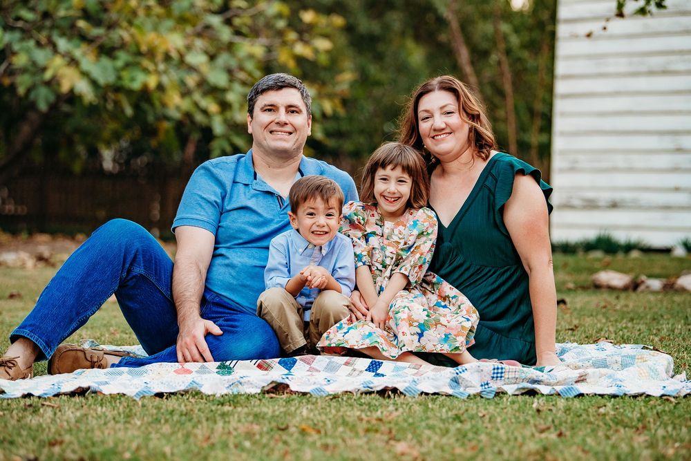 A family of four sits together on a colorful quilt on green grass, smiling for a portrait.