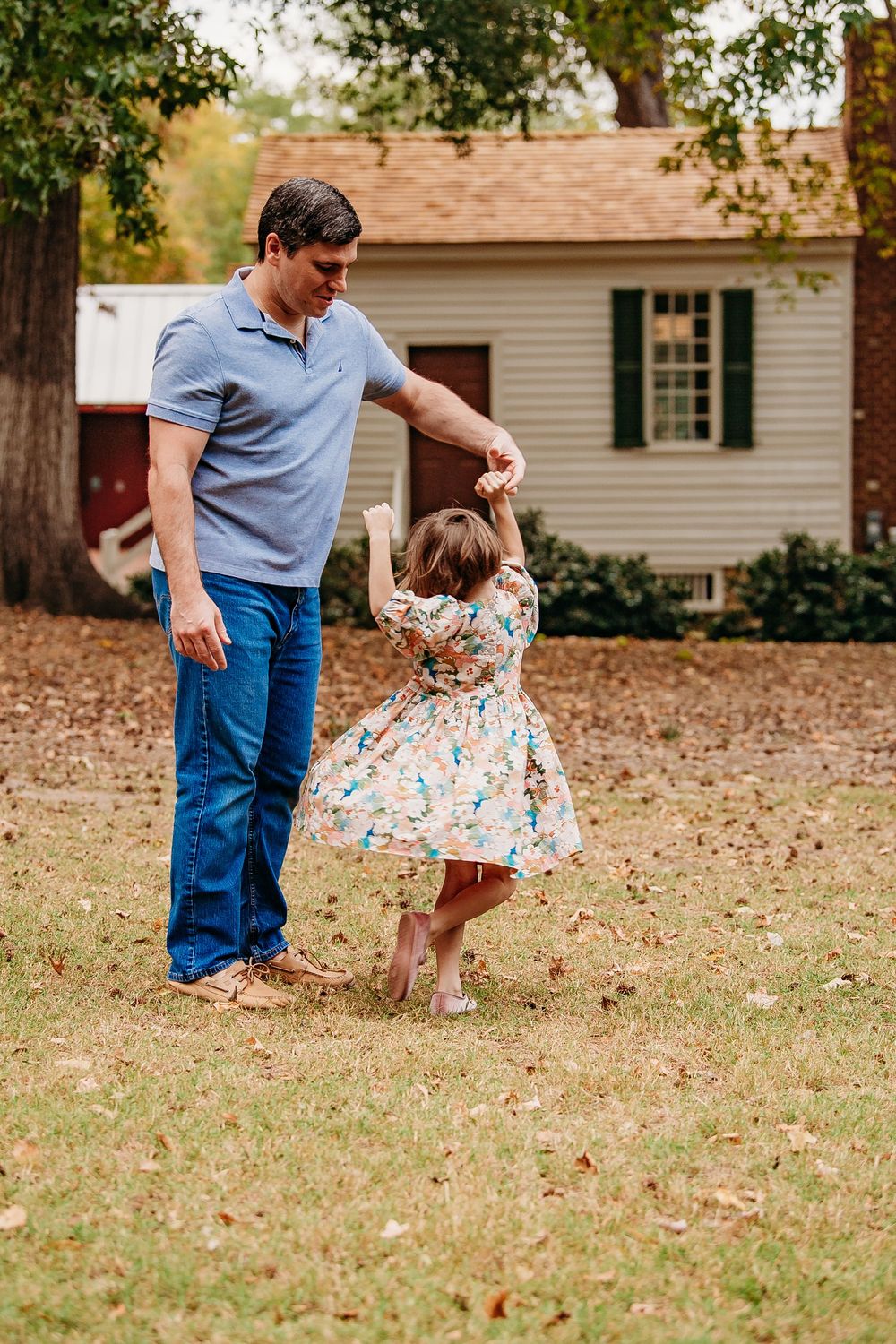 Father spinning his young daughter in a floral dress in a leaf-covered backyard.