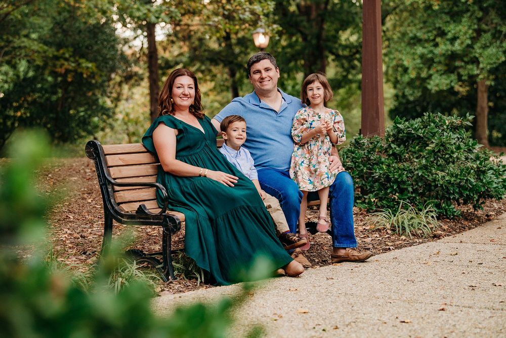 Family of four sitting together on a park bench surrounded by lush green trees.