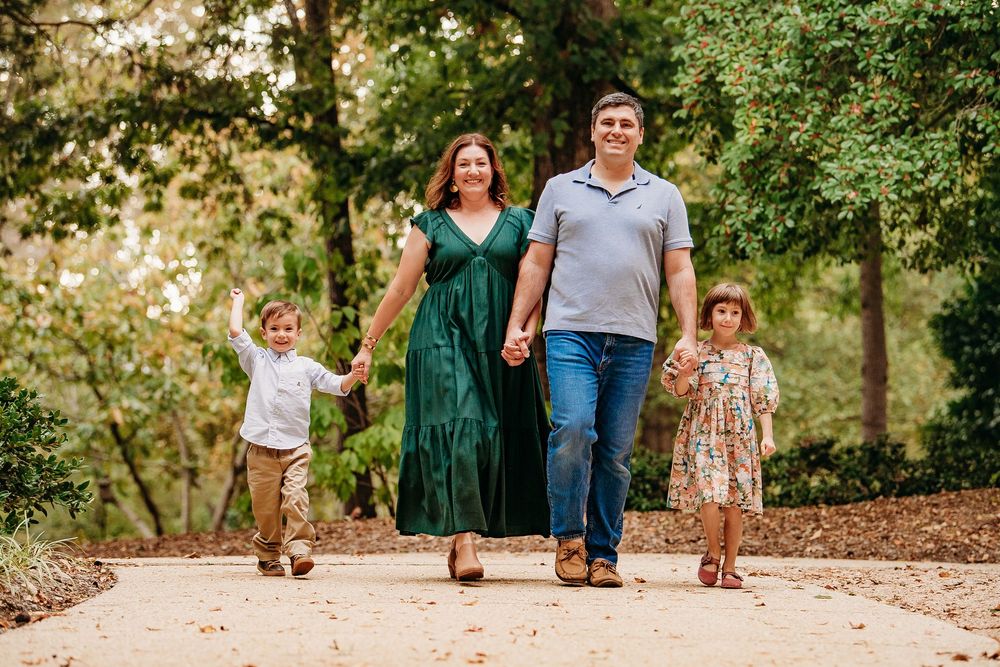 Happy family of four walking hand in hand along a tree-lined park pathway.