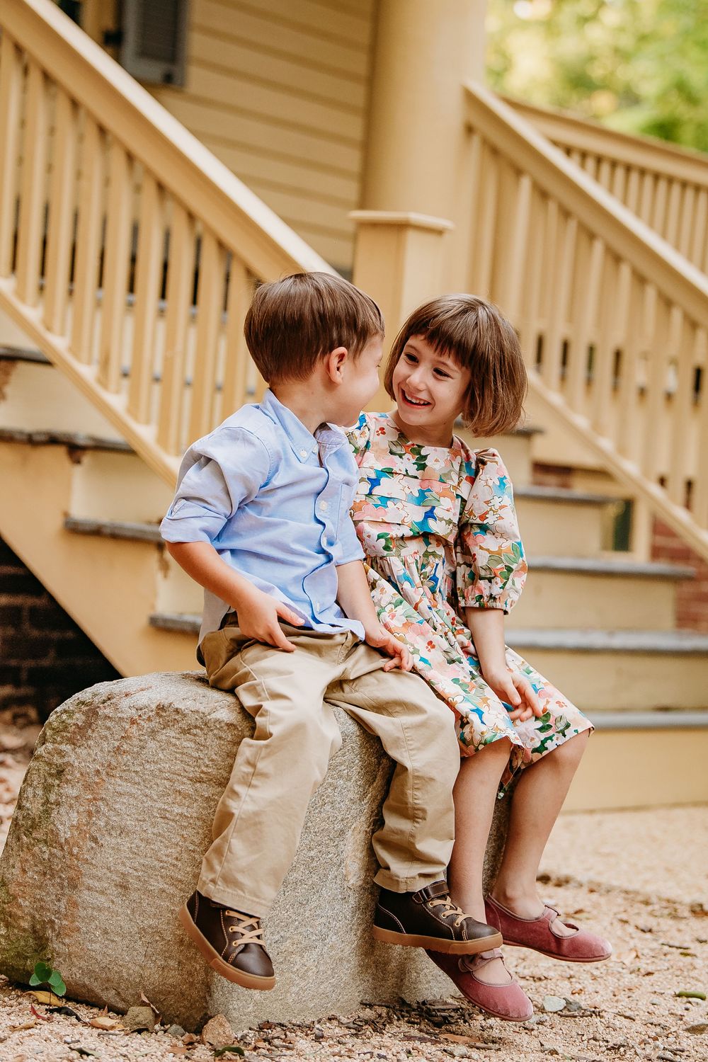 Two young children sit together on a stone step, smiling and sharing a sweet moment outside a home.