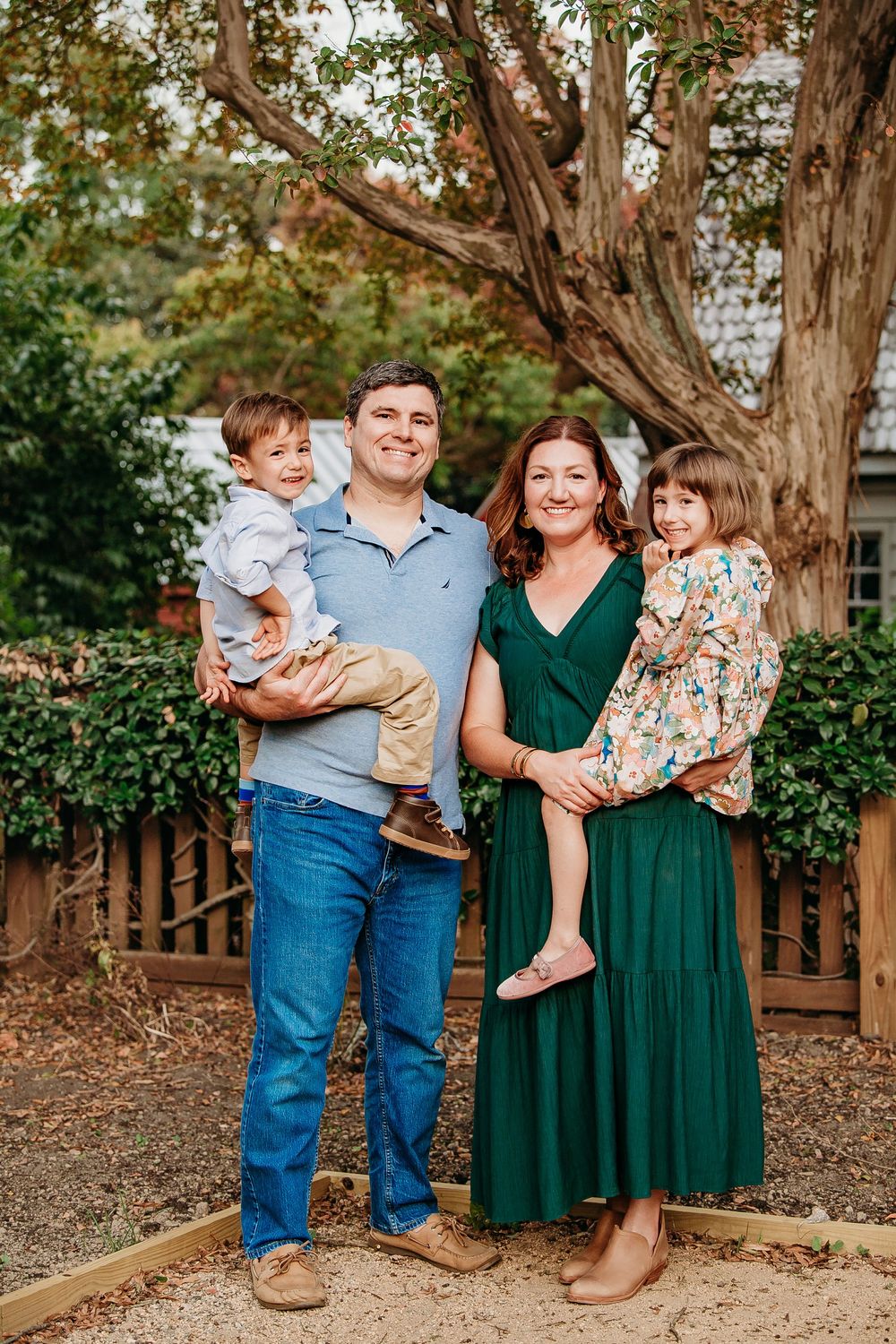 A happy family of four poses outdoors, parents holding their two young children near a large tree.