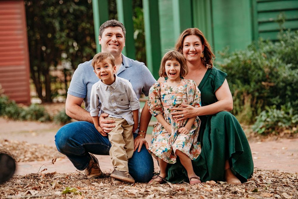 A smiling family of four sits together outdoors on a sunny day, posing for a portrait photo.