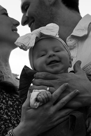 Smiling parents hold their laughing baby girl wearing a bow headband in black and white photo.