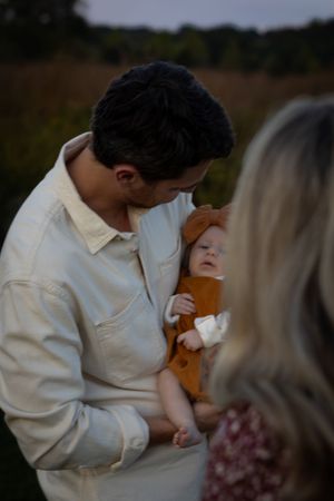 Father in cream shirt tenderly kisses his infant daughter dressed in rust-colored outfit outdoors.