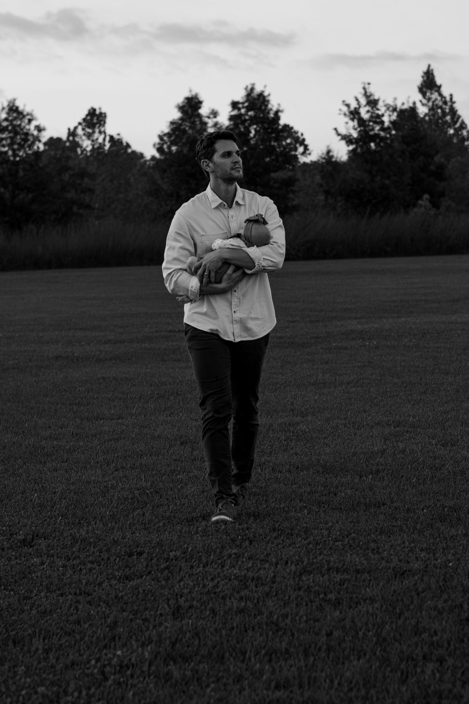 Man in white shirt walks across open grass field carrying sleeping newborn baby at dusk.