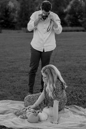 Father photographs mother and baby on blanket with pumpkin during outdoor fall family session.