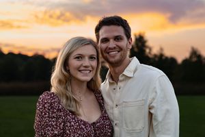Smiling couple poses together in front of a vibrant golden sunset over a green field.