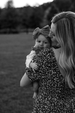 Mother in floral dress gently kisses her baby girl wearing a bow headband in black and white.