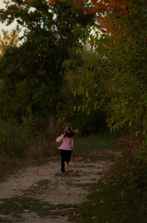 A young girl in a pink jacket runs down a leaf-covered forest path surrounded by autumn foliage.