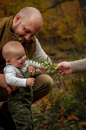 Smiling bald father holding baby in autumn forest while partner offers a green leaf.