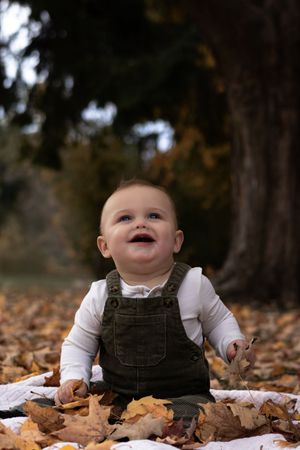 Adorable smiling baby in green overalls sitting on fallen autumn leaves in a park.