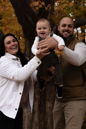 Happy family with laughing baby held up between smiling parents in autumn park.