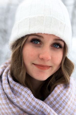 Young woman with blue eyes wearing white knit beanie and plaid scarf in snowy winter setting.