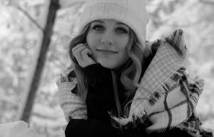 Black and white portrait of blonde woman in white beanie and plaid scarf in winter snow.