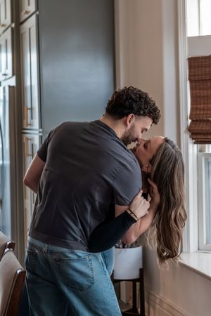 Couple sharing a romantic kiss near a window in a cozy indoor setting.