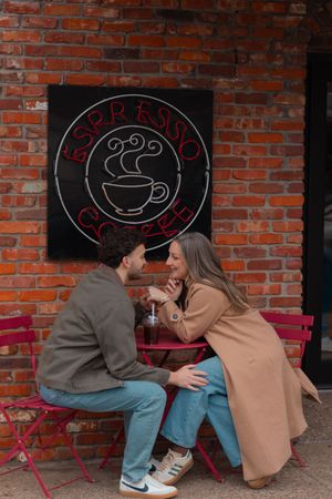 Romantic couple sitting at outdoor café table in front of espresso coffee neon sign on brick wall.