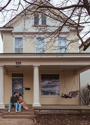 Couple sitting on porch steps of charming yellow Victorian house numbered 233 in winter.