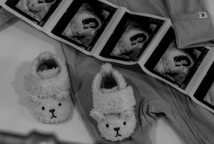 Black and white photo of cute bear baby booties beside ultrasound photos on a soft blanket.