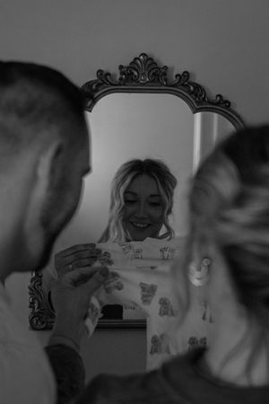 Black and white reflection of a smiling blonde woman looking at a pregnancy test in an ornate mirror.