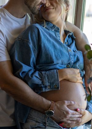 Couple tenderly embracing as man holds pregnant woman's bare baby bump near a bright window.