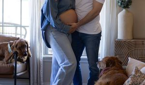 Pregnant couple embracing near a bright window while two dachshund dogs relax on the couch.